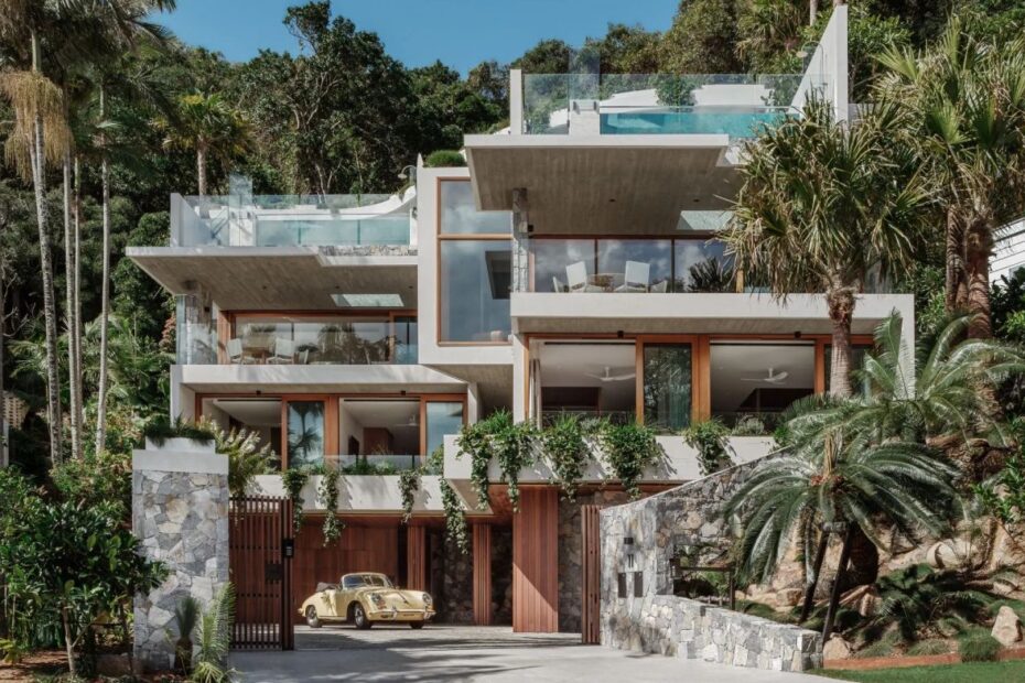 Modern multi-level tropical villa with glass balconies, stone walls, and lush palm trees; a yellow vintage car in the driveway.