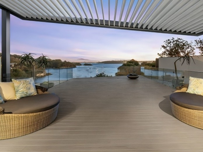 Rooftop terrace with curved wicker lounge beds and cushions, glass railing, and a water view at sunset.