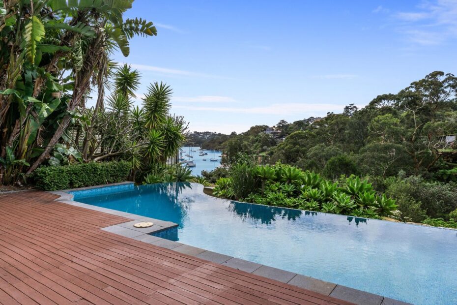 Infinity pool with a wooden deck and lush tropical plants, overlooking a harbor with boats and tree-covered hills under a blue sky