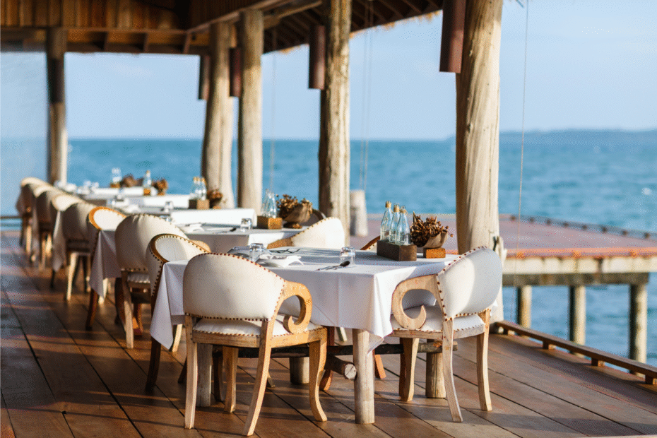 seaside open-air restaurant with white-tablecloth tables and cream chairs along a wooden deck by the water, ready for guests