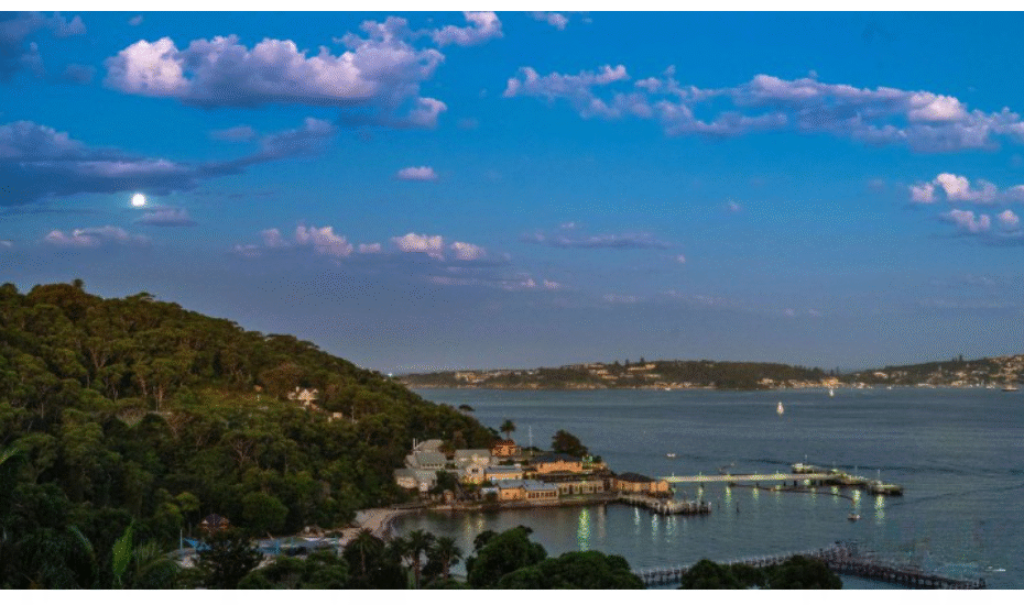 coastal town by a calm bay with a wooden pier, houses along the shore, and a forested hill under a blue sky with clouds.