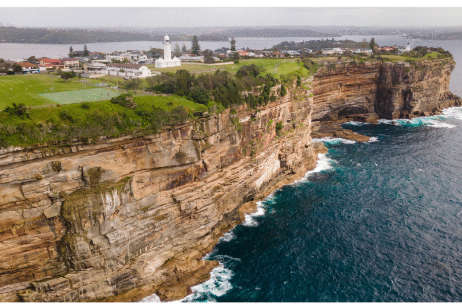 coastal cliff-top park with a white lighthouse, adjacent houses, and a tennis court overlooking the ocean.