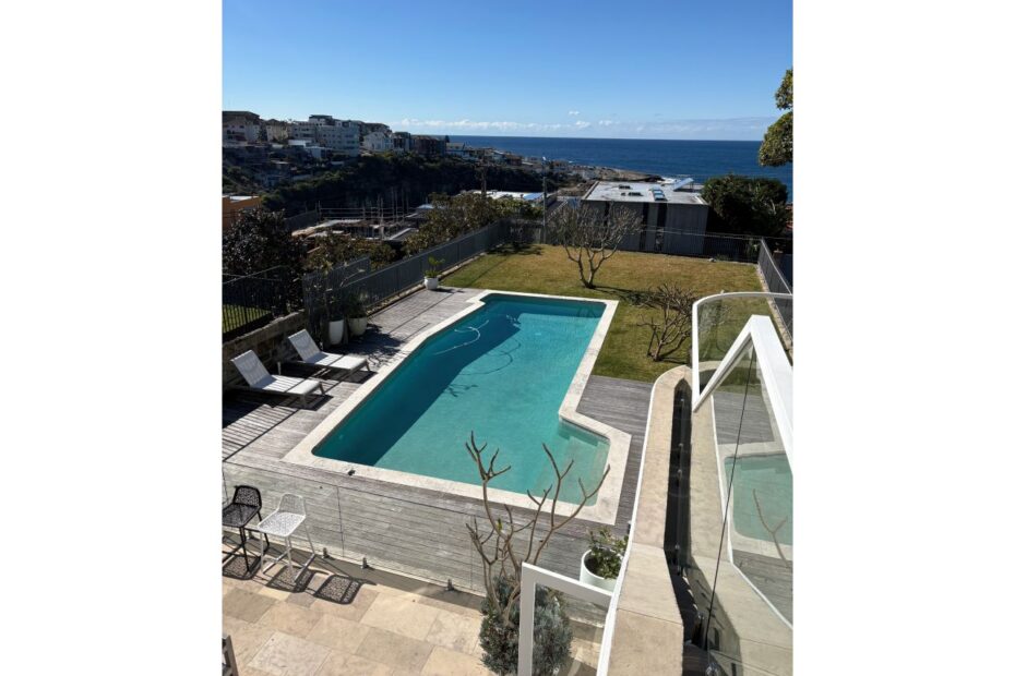Backyard pool area with a wooden deck, white lounge chairs, and an ocean view beyond a fence.